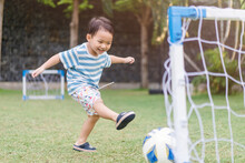 Asian Toddler Boy Playing Soccer Or Football In Football Field Park Outdoor.3 Years Old Asian Boy Kicking Football On The Grass Field.Little Boy Shooting At Goal, Winner, Sport Activity In Sport Club.