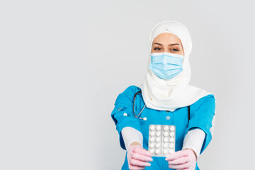 Wall Mural - friendly Muslim doctor or nurse in a hijab, mask, gloves offering a pill to the patient on a gray background