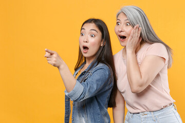 Wall Mural - Shocked family asian women girls gray-haired mother and brunette daughter in casual clothes posing pointing index finger aside put hand on cheek isolated on yellow color background studio portrait.