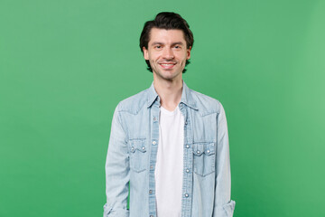 Handsome smiling cheerful young brunet man 20s wearing casual clothes white t-shirt denim shirt posing standing and looking camera isolated on green color wall background, studio portrait.