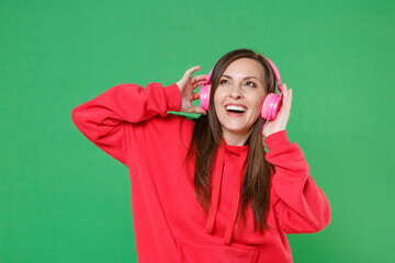 Wall Mural - Cheerful young brunette woman 20s wearing bright red casual streetwear hoodie posing listening music with headphones looking up put hands on head isolated on green color background studio portrait.