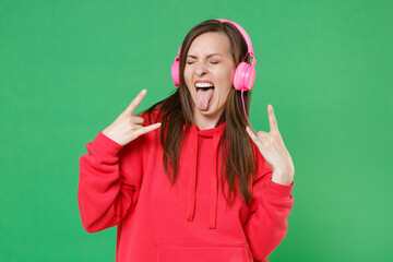 Wall Mural - Funny young brunette woman 20s wearing bright red casual streetwear hoodie posing listening music with headphones depicting heavy metal rock sign isolated on green color background studio portrait.