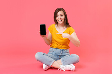 Wall Mural - Full length portrait of excited young woman in yellow t-shirt posing sit on floor pointing index finger on mobile phone with blank empty screen mock up copy space isolated on pink background studio.