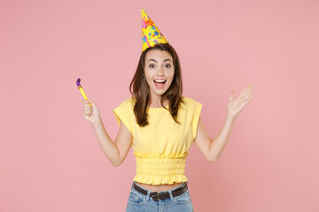 Wall Mural - Excited surprised young brunette woman 20s wearing yellow casual t-shirt birthday hat posing standing celebrating holding pipe spreading hands isolated on pastel pink color background studio portrait.