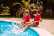 © Dragica - Two beautiful caucasian boys in red T-shirts are sitting on the edge of the pool and spraying water with their feet. Children at the pool by the palm trees
