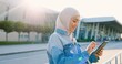 Young muslim pretty joyful woman in traditional hijab using tablet device at street. Urban landscape. Beautiful happy islamic female tapping and scrolling on gadget. Outdoors.