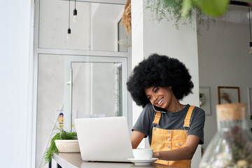 Happy young adult hipster African American woman, female student with afro curly hair holding smartphone talking with friend on the phone or making business call on cellphone sitting at cafe table.