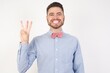 © Roquillo - European man dressed in formal shirt and bow tie poses against white studio background showing and pointing up with fingers number three while smiling confident and happy.