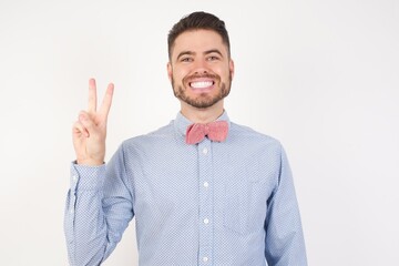 European man dressed in formal shirt and bow tie poses against white studio background showing and pointing up with fingers number two while smiling confident and happy.