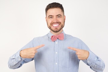 Wall Mural - Horizontal shot of European man dressed in formal shirt and bow tie poses against white background points at his body, being in good mood after going shopping and making successful purchases
