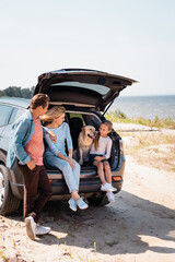 Wall Mural - Selective focus of parents looking at daughter with golden retriever in car trunk on beach