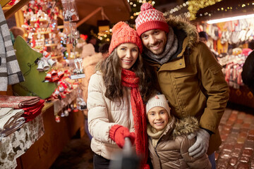 Sticker - family, winter holidays and technology concept - happy mother, father and little daughter taking picture by selfie stick at christmas market on town hall square in tallinn, estonia