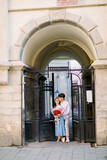 Fototapeta Na drzwi - City outdoor portrait of happy young couple in love, posing near pod building with beautiful vintage gate. Pretty girl in blue dress holds bouquet of red flowers, handsome guy kisses her