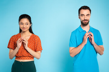 Wall Mural - sly asian woman in red blouse and tricky man in polo t-shirt gesturing with joined fingers on blue