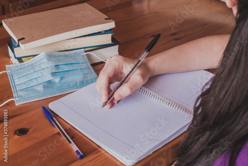 Una chica sujeta un bolígrafo mientras estudia en una mesa con libros, una mascarilla azul y un cuaderno en blanco