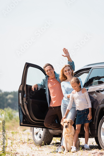 Selective focus of man waving hand near family with golden retriever and auto outdoors