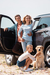 Wall Mural - Selective focus of man embracing wife near daughter, golden retriever and auto outdoors