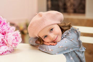 Happy little girl with curly hair in a dress and in a beret sits at a table with a bouquet of pink peonies in a street vintage cafe. Childhood concept. French style. Birthday. Mothers Day. Celebration