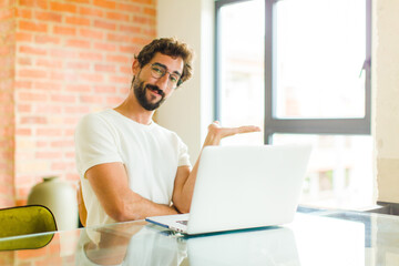 young bearded man with a laptop smiling, feeling confident, successful and happy, showing concept or idea on copy space on the side