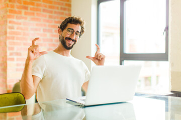 Wall Mural - young bearded man with a laptop framing or outlining own smile with both hands, looking positive and happy, wellness concept