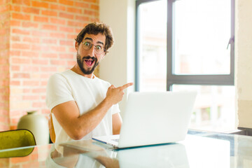 Wall Mural - young bearded man with a laptop looking excited and surprised pointing to the side and upwards to copy space