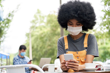 Wall Mural - African American young hipster gen z female with Afro hair wearing face mask holding smart phone using mobile apps sitting at table in outdoor cafe. Social distancing everyday life and tech concept