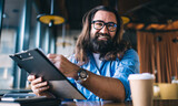 Portrait of cheerful bearded male owner of small business enjoying work in cafe satisfied with finance income, prosperous mature man in spectacles holding planner with document looking at camera