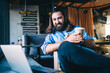 © BullRun - Positive caucasian bearded mature man enjoying rest on free time in cafe interior watching movie on laptop computer holding coffee cup, smiling 40 years male enjoying reading news on leisure