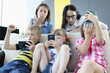 Three children are sitting on the couch with smartphones in their hands, playing online games from back of couch, two women are standing and looking frightened at phone screen. Child protection from