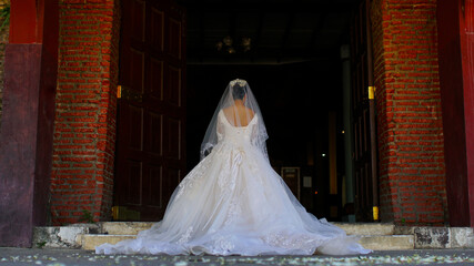 Full length side view of one beautiful young bride in long white wedding dress and veil standing in forest holding bouquet outdoor on natural background