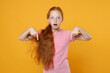 Shocked little ginger redhead kid girl 12-13 years old in pink casual t-shirt posing pointing index fingers down on mock up copy space isolated on yellow color background children studio portrait.