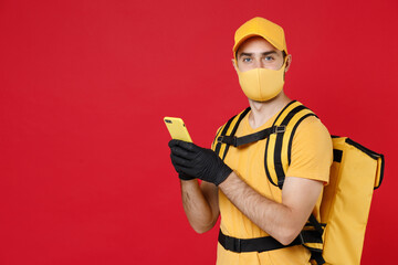 Wall Mural - Delivery employee man in yellow cap face mask gloves t-shirt uniform thermal food bag backpack, hold cellphone, work courier service during quarantine covid-19 virus isolated on red background studio.