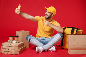 Wall Mural - Delivery employee man guy male in yellow cap t-shirt sit at thermal takeaway food bag backpack pizza boxes cups hold mobile cell phone work courier service during covid-19 isolated on red background.