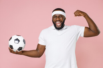 Cheerful young bearded african american fitness sports man football player in headband t-shirt playing soccer hold ball showing biceps muscles isolated on pastel pink color background studio portrait.