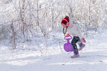 The baby is playing in a snow-covered glade. Concept of active children's recreation. A cheerful little girl runs with an iceboat in her hand. A child runs on a hill in the winter forest.