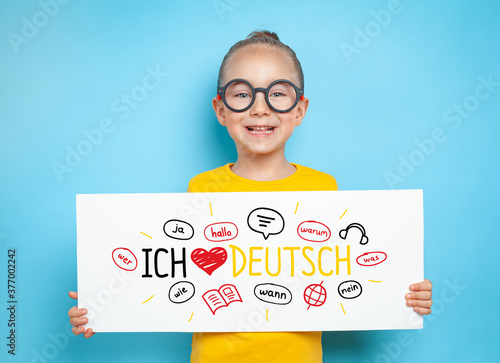Photo Smiling beautiful cute little girl holding a white paper with ich liebe deutsch text and illustrations while looking at the camera