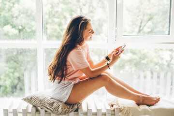 Attractive young woman in pink t-shirt with cup of coffee surfs internet with modern mobile phone sitting on white bench near wide window in light room