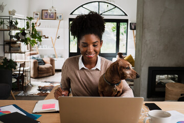 Smiling female student on video call catching up with friends from university, sitting with puppy studying in modern lounge.