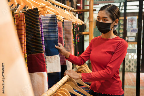 Plakat Young travel shopping woman choosing unique Thai native clothes at village shop.