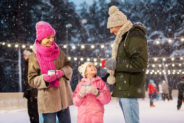 Wall Mural - christmas, family and leisure concept - happy mother, father and daughter drinking hot tea at outdoor skating rink in winter