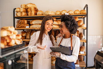 Attractive women selling fresh pastry and loaves in bread section and smiling. Portrait of two beautiful young bakers at the bakery. Business owner talking to worker at a bakery