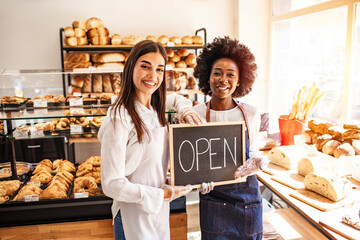 Portrait of two young entrepreneurs standing in the bakery shop with OPEN sign. Two cheerful small business owners smiling and looking at camera while standing with open sign board.