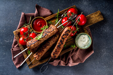 Shish kebab on skewers, from minced beef meat. Lula kebab, traditional Caucasian recipe. On black concrete background, with sauces, tomatoes and veggie salad top view copy space