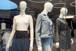 three plastic female white mannequins dressed in dresses and denim clothing through a glass window in a store in a shopping center