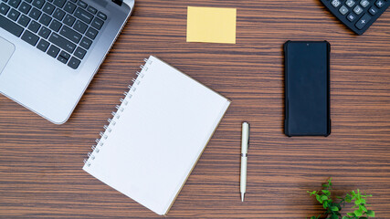 An office table working enviroment. Notepad, sticky note, pen plant, calculator and a lap top on a brown striped zebrawood design table top. Must have objects while working from home during Covid-19