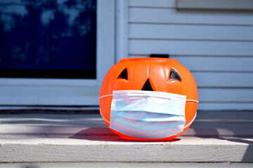 Coronavirus Halloween Pumpkin. Halloween jack o lantern wearing a covid-19 face mask and placed outside a home at daytime. House, front door, and window visible in background.