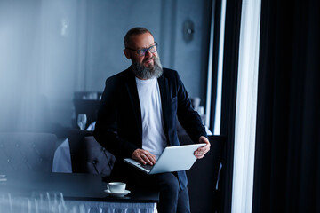 An adult businessman with a beard in glasses sits with a laptop on his lap and works. The director makes a schedule for the workflow