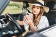 © Miljan Živković - Front view on beautiful caucasian woman in hat and white dress using mobile phone texting while driving her car in nature in bright summer or autumn day - travel vacation concept yellow filter