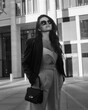 © Dmitry Tsvetkov - Young stylish woman in white overall, sneakers, black leather jacket and sunglasses holding small handbag and standing at city street. Fashion style outdoor portrait