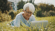 old gray haired woman using laptop in the nature while lying on the grass. High quality photo